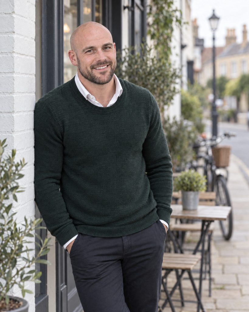 A fit financial-planning professional smiles outside a south England cafe in a relaxed, friendly portrait.